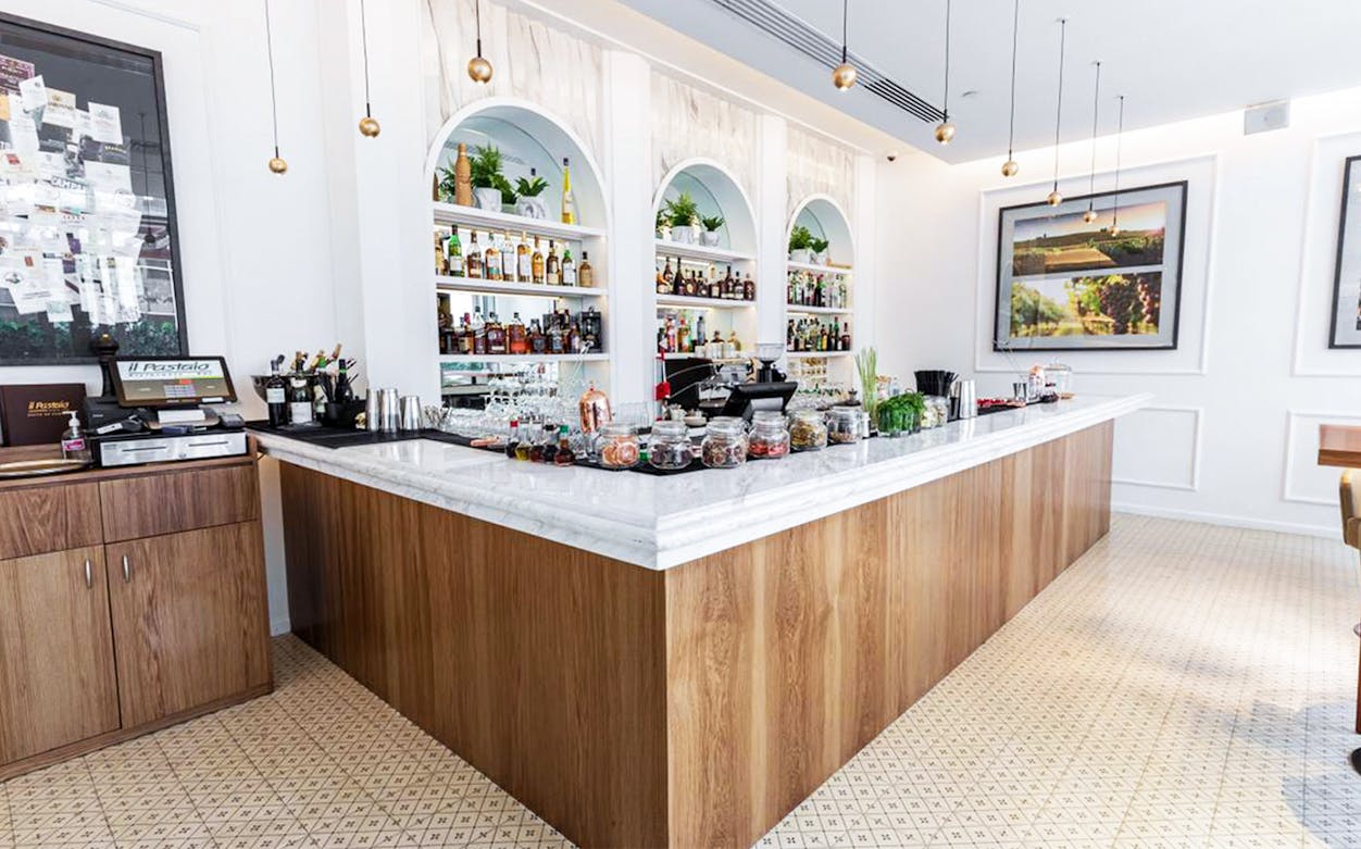 Bar area at Il Pastaio Restaurant, featuring a marble countertop and shelves with bottles, part of La Perle dinner package.