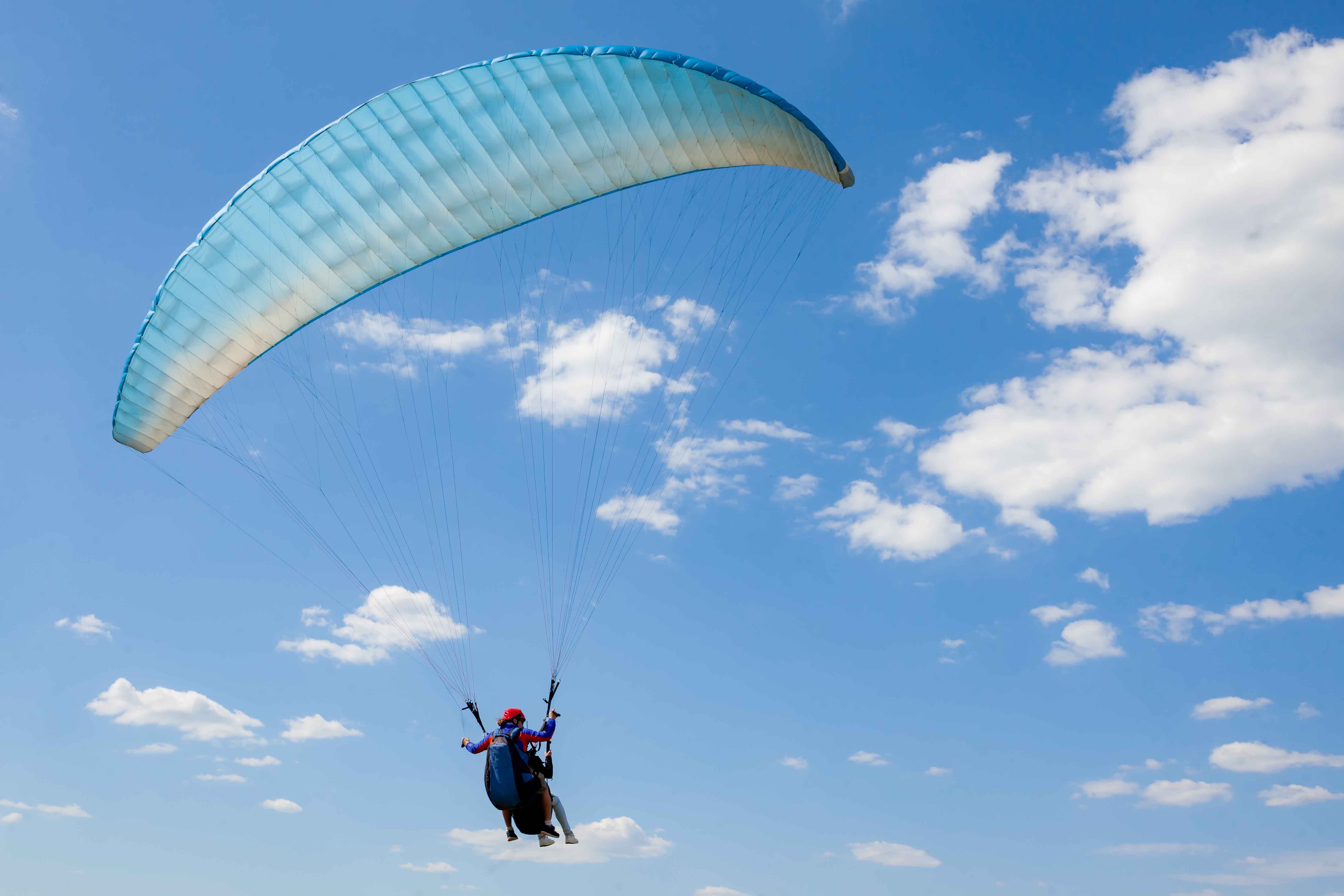 Paragliding tandem flight against a clear blue sky with scattered clouds.