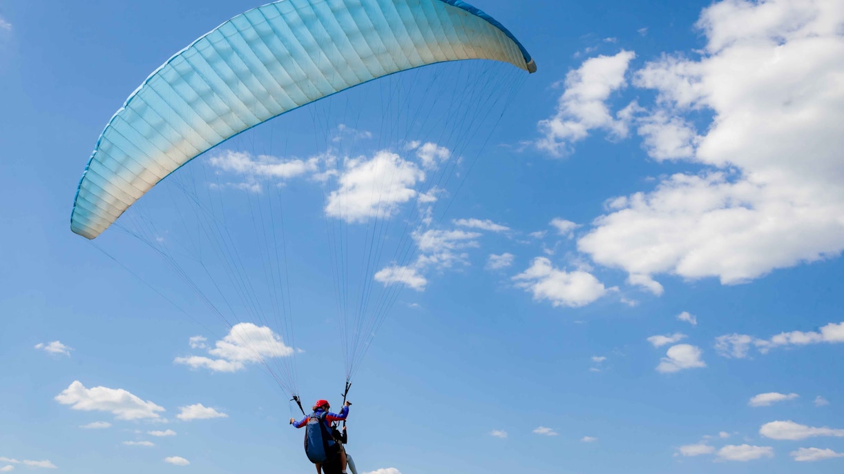Paragliding tandem flight against a clear blue sky with scattered clouds.