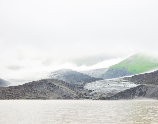 Falljokull glacier shrouded in fog with rocky terrain and green hills, Iceland.