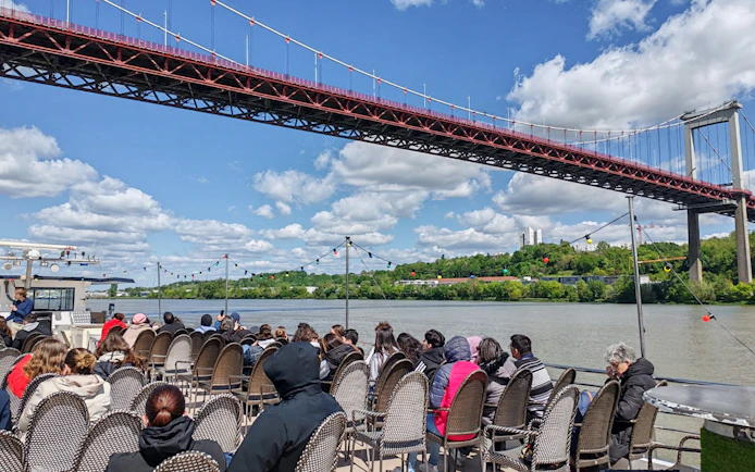Tourists seated on a boat during a Bordeaux sightseeing river cruise under a large bridge.