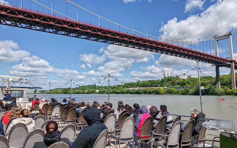 Tourists seated on a boat during a Bordeaux sightseeing river cruise under a large bridge.