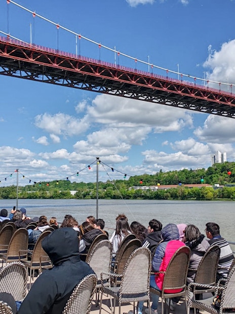 Tourists seated on a boat during a Bordeaux sightseeing river cruise under a large bridge.