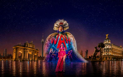 Performer in vibrant costume at WAH Show with illuminated cityscape backdrop.