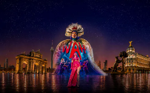 Performer in vibrant costume at WAH Show with illuminated cityscape backdrop.