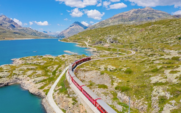 Bernina train traveling through Bernina Pass with lake and mountains in summer.
