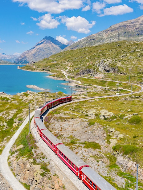 Bernina train traveling through Bernina Pass with lake and mountains in summer.