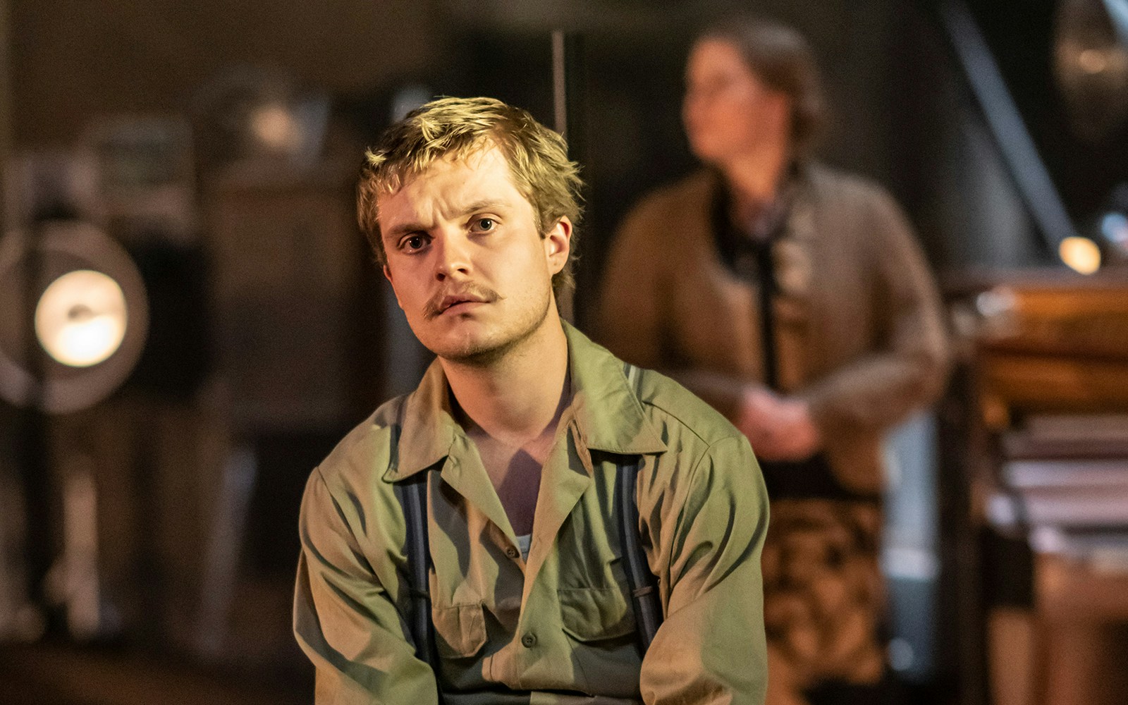 Actor in a scene from "The Glass Menagerie" play, with a blurred figure in the background.