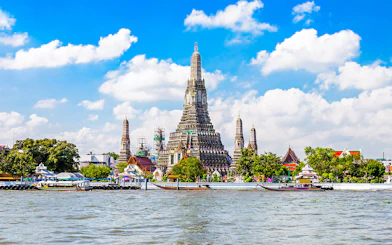Wat Arun temple along Chao Phraya River in Bangkok with boats passing by.