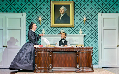 Theatrical scene with actors in period costumes, ornate desk, and portrait backdrop.