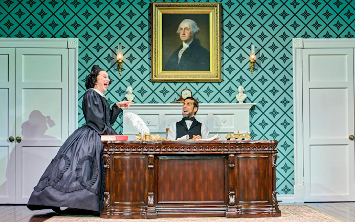 Theatrical scene with actors in period costumes, ornate desk, and portrait backdrop.