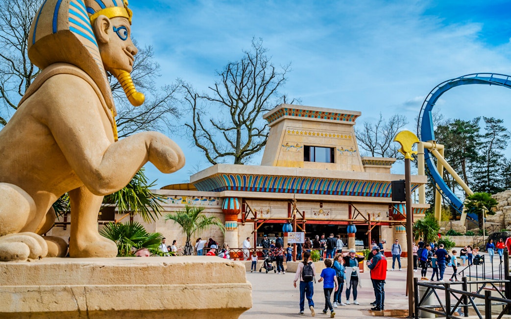 Roller coaster loop and Egyptian-themed entrance at Parc Asterix, France, with tourists exploring.