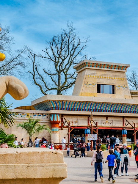 Roller coaster loop and Egyptian-themed entrance at Parc Asterix, France, with tourists exploring.