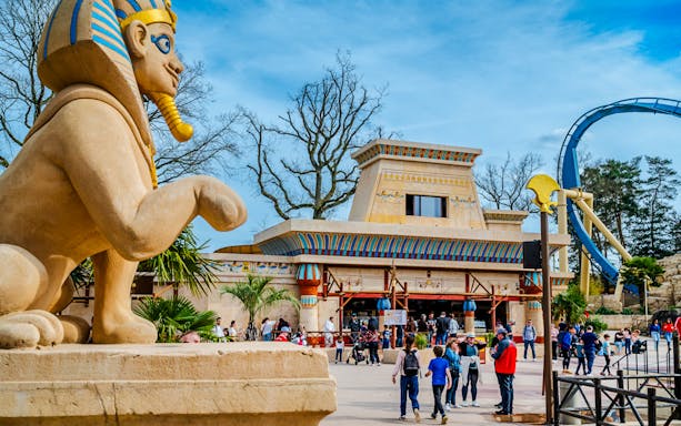 Roller coaster loop and Egyptian-themed entrance at Parc Asterix, France, with tourists exploring.