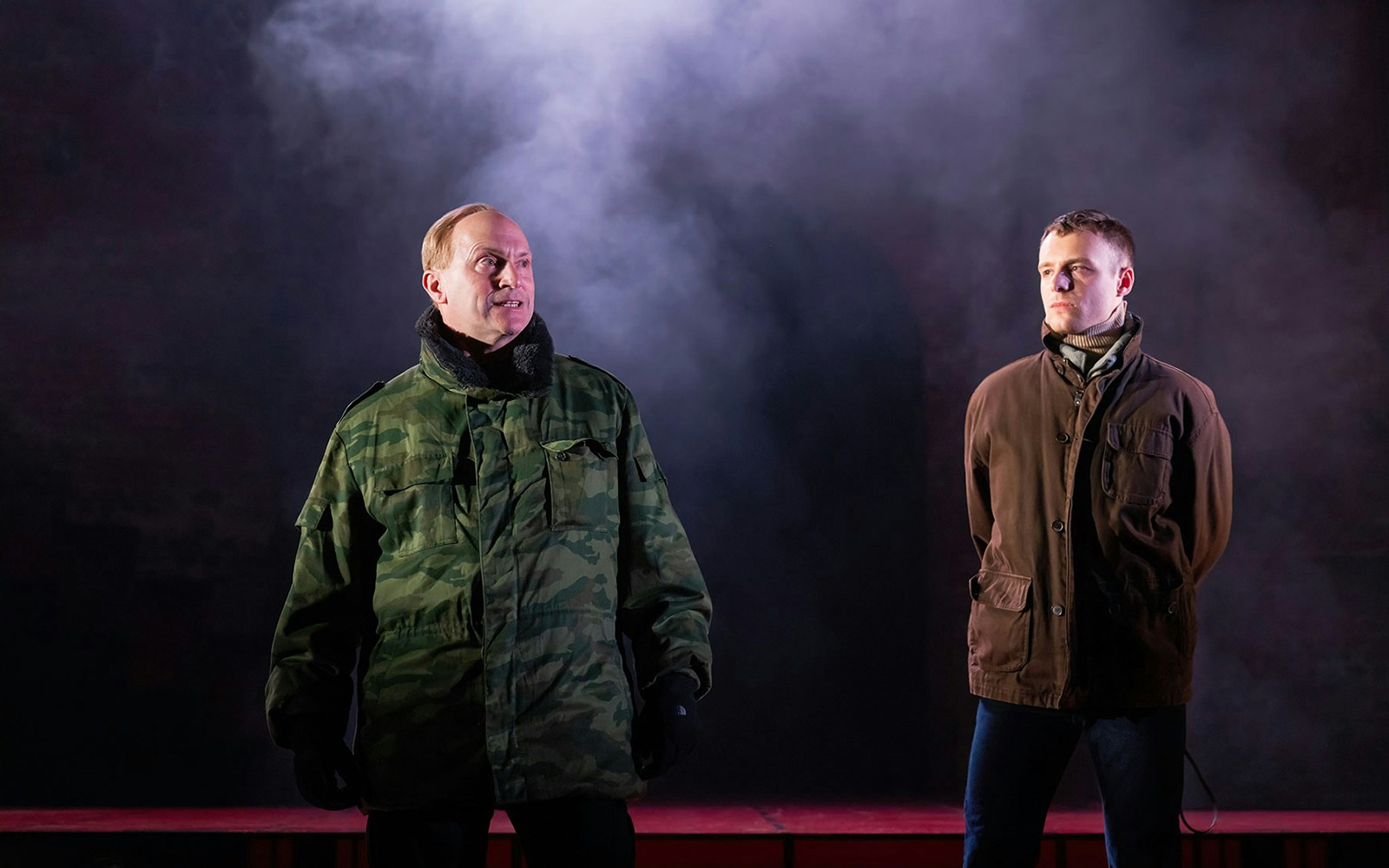 Two men in military and casual jackets stand on stage with smoke backdrop, related to Patriots.