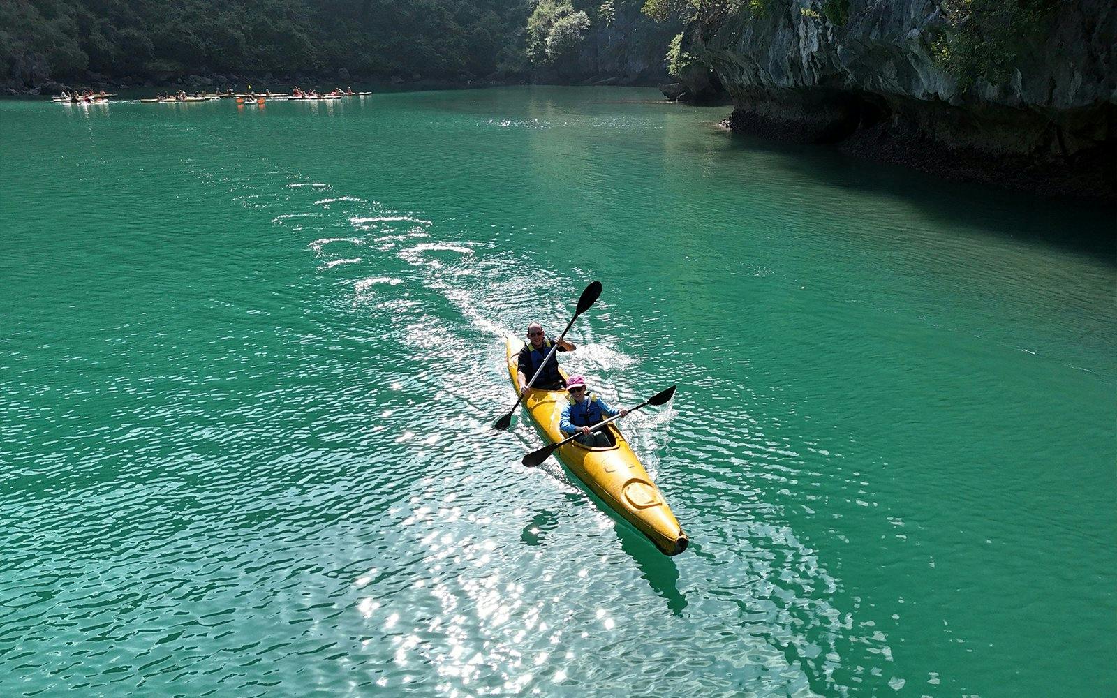 Kayakers paddling in a yellow kayak on emerald waters near rocky cliffs.