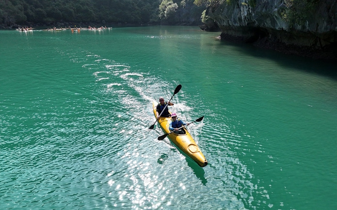 Kayakers paddling in a yellow kayak on emerald waters near rocky cliffs.
