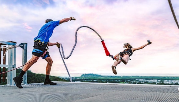 Person bungee jumping from rooftop with instructor, scenic city view in background.
