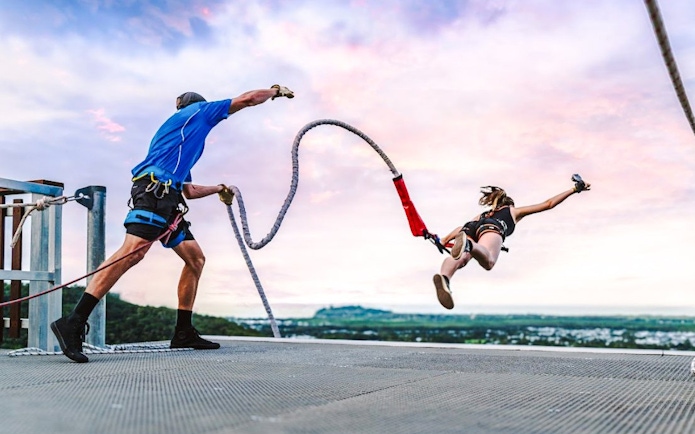 Person bungee jumping from rooftop with instructor, scenic city view in background.