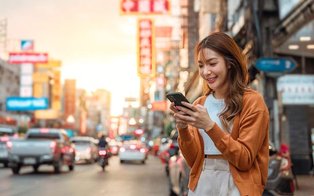 Young woman using smartphone on busy Asian city street at sunset.
