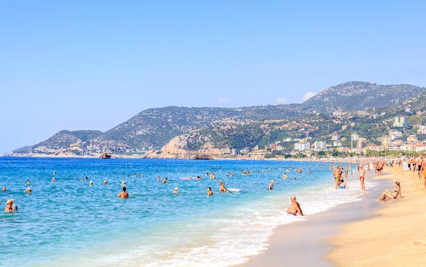 Beachgoers enjoying the sea and sun in Alanya, Turkey, with scenic mountains in the background.