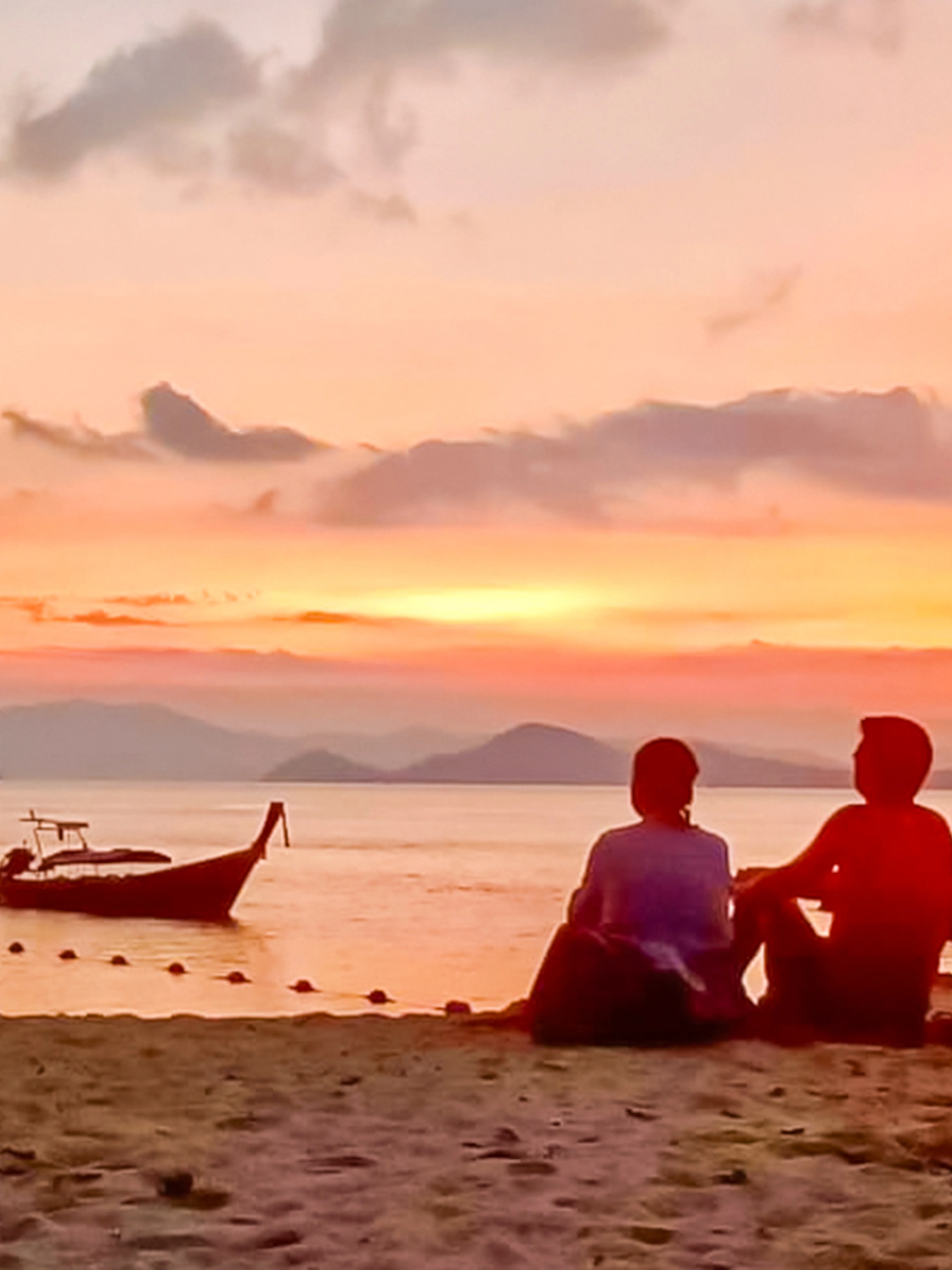 Couple sitting on Krabi beach at sunset with a boat and torch nearby.