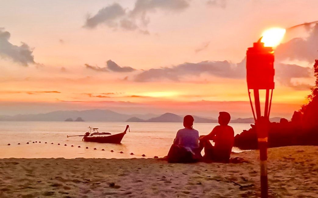 Couple sitting on Krabi beach at sunset with a boat and torch nearby.