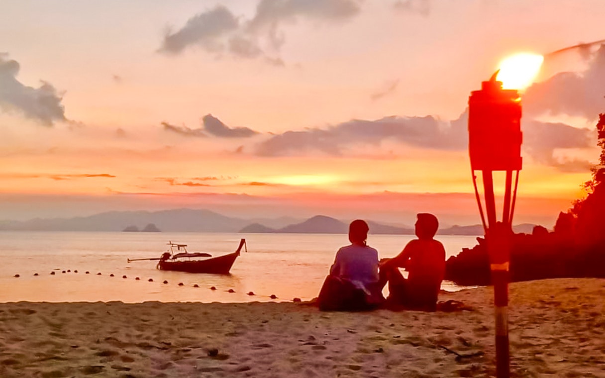 Couple sitting on Krabi beach at sunset with a boat and torch nearby.