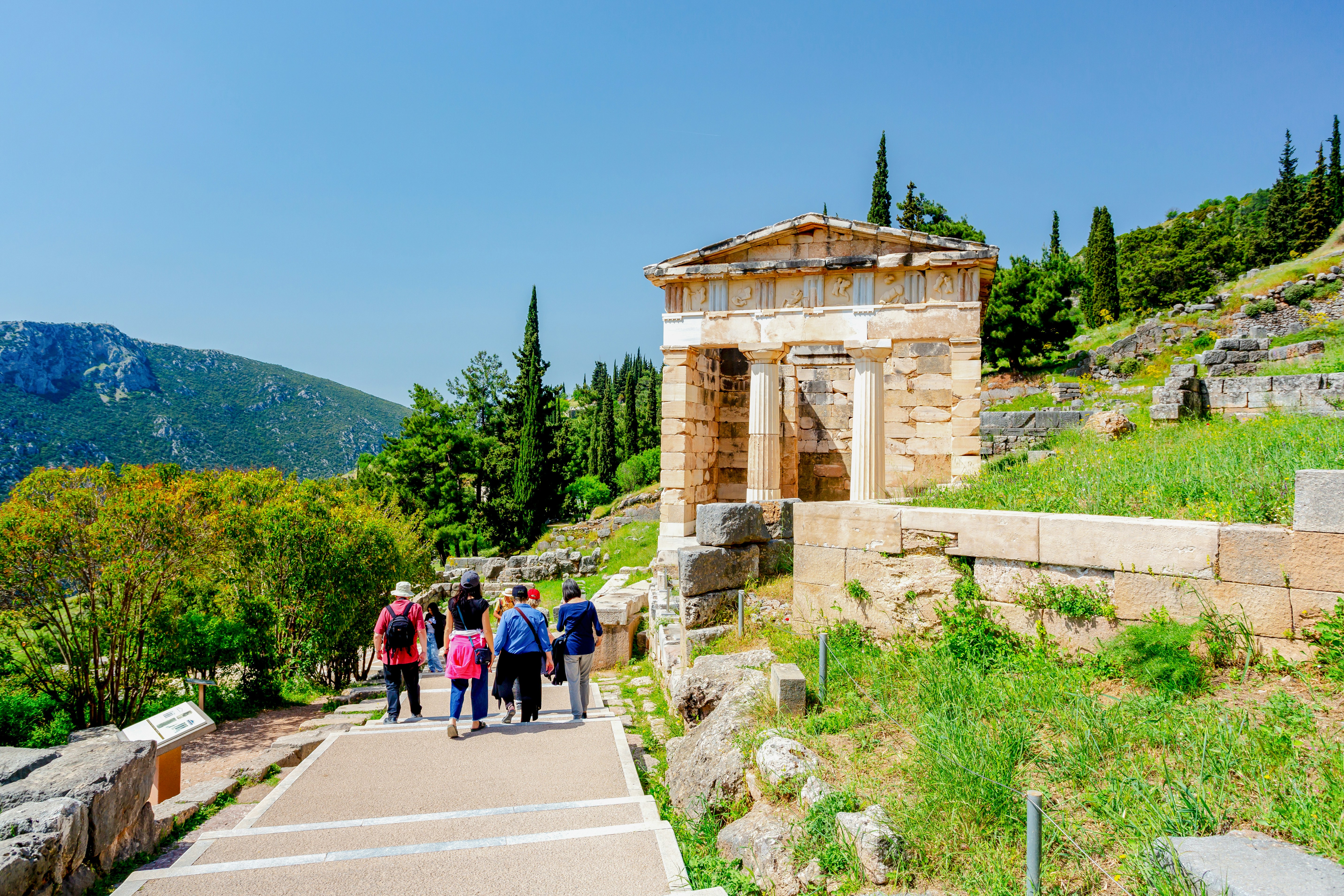 Tourists walking towards the Athenian Treasury at Delphi, Greece, surrounded by greenery.