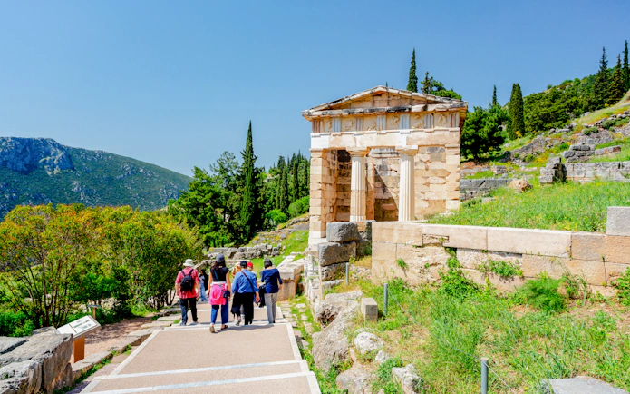Tourists walking towards the Athenian Treasury at Delphi, Greece, surrounded by greenery.
