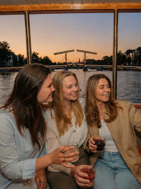 Three people enjoying drinks on a canal cruise in Amsterdam at sunset with a view of the Skinny Bridge.