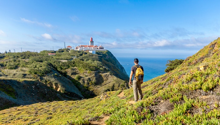 Traveler hiking towards Cabo da Roca lighthouse with Atlantic Ocean in the background.