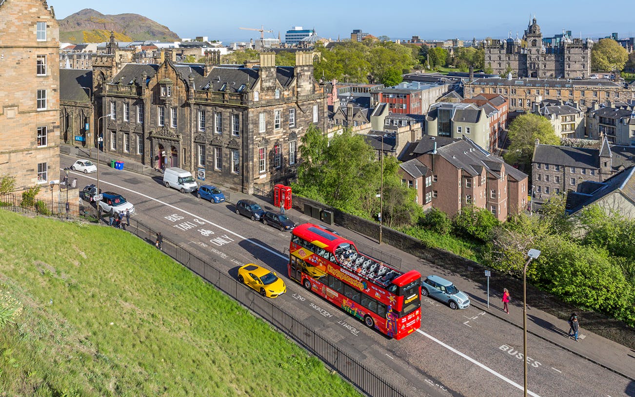 HOHO bus on street near Edinburgh Castle with cityscape in background.