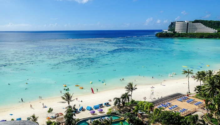 Tumon Bay Beach in Guam with clear blue waters and palm trees lining the sandy shore.