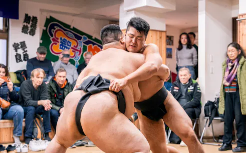Sumo wrestlers grappling during a match in a Tokyo dojo, with spectators watching closely.