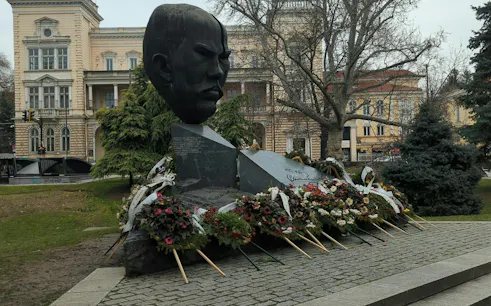 Bust monument with floral tributes in Sofia, part of Sofia Obscura Secrets exploration game.