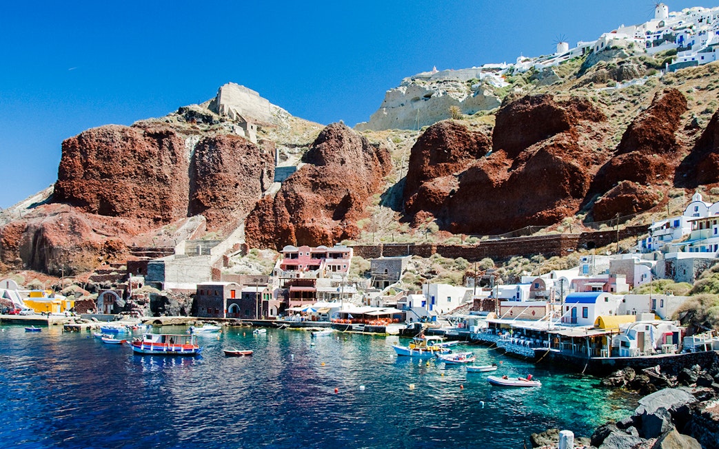 Boats docked at Amoudi port below Oia caldera, Santorini, with red cliffs and white buildings.