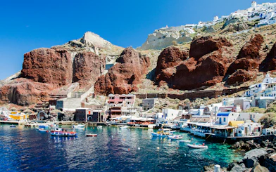 Boats docked at Amoudi port below Oia caldera, Santorini, with red cliffs and white buildings.
