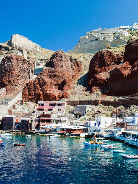 Boats docked at Amoudi port below Oia caldera, Santorini, with red cliffs and white buildings.