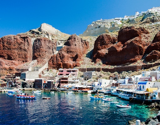Boats docked at Amoudi port below Oia caldera, Santorini, with red cliffs and white buildings.