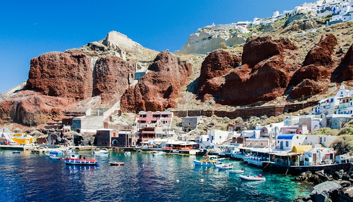 Boats docked at Amoudi port below Oia caldera, Santorini, with red cliffs and white buildings.