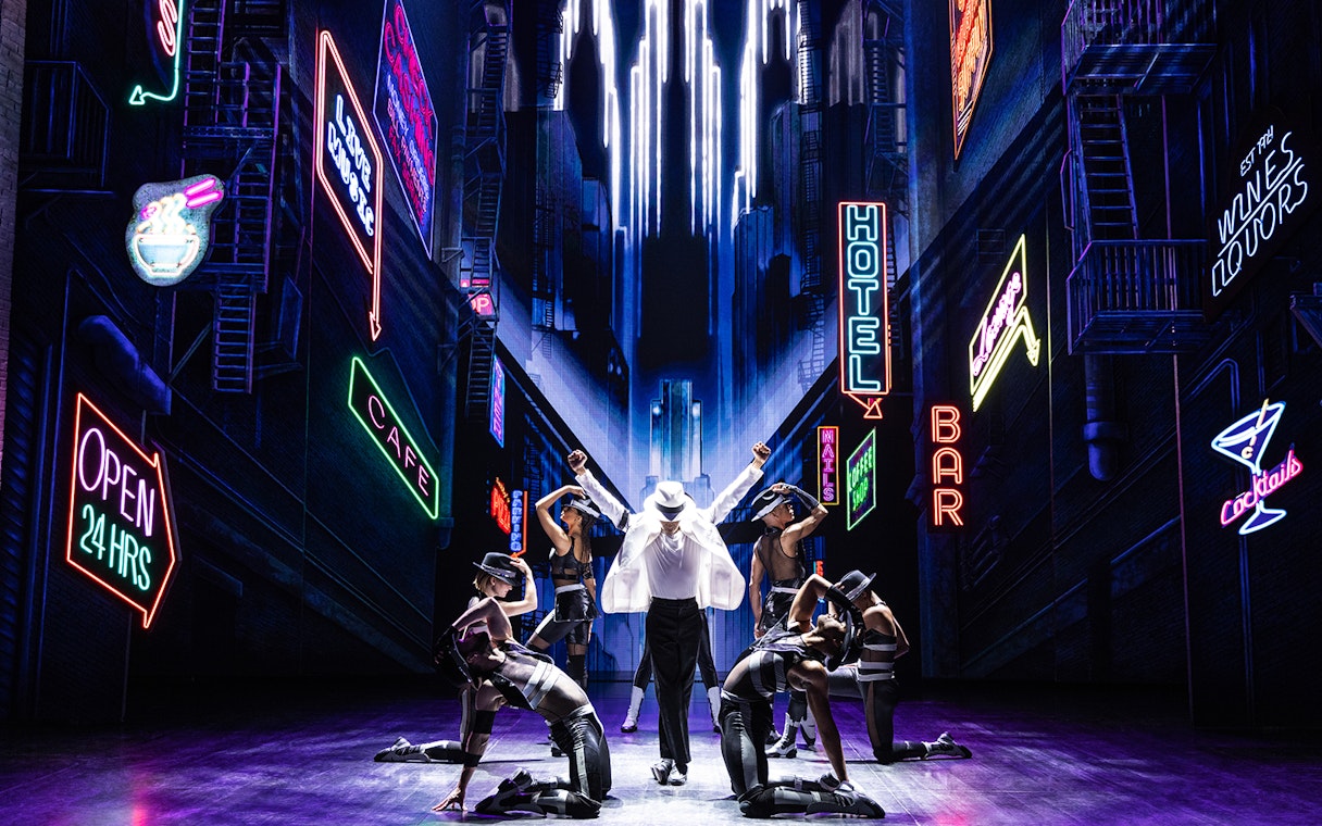 Performer in white suit and dancers on stage with neon cityscape backdrop at MJ Live Tribute Concert.