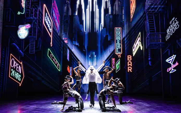 Performer in white suit and dancers on stage with neon cityscape backdrop at MJ Live Tribute Concert.