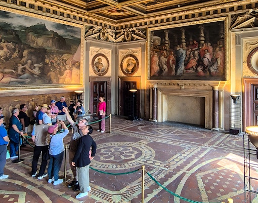 Visitors exploring the ornate interior of Palazzo Vecchio, Florence, with large frescoes on the walls.