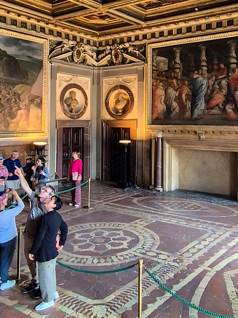 Visitors exploring the ornate interior of Palazzo Vecchio, Florence, with large frescoes on the walls.
