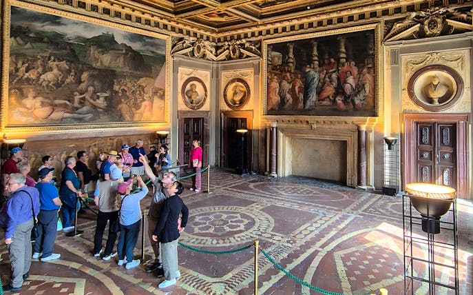Visitors exploring the ornate interior of Palazzo Vecchio, Florence, with large frescoes on the walls.