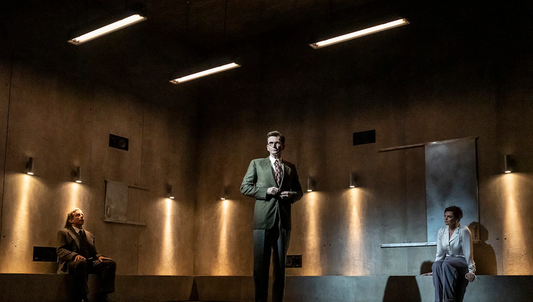 Three people in a dimly lit room, two seated and one standing, during a theatrical performance.