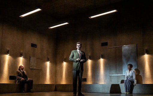 Three people in a dimly lit room, two seated and one standing, during a theatrical performance.