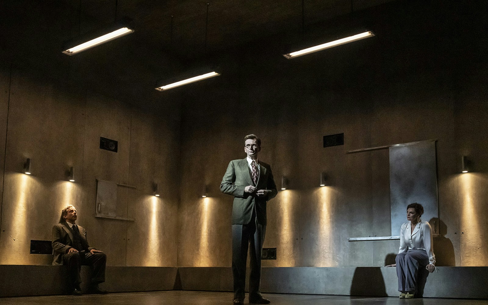 Three people in a dimly lit room, two seated and one standing, during a theatrical performance.