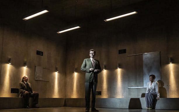 Three people in a dimly lit room, two seated and one standing, during a theatrical performance.